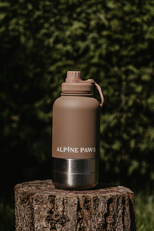 Brown water bottle with 'Alpine Paws' branding on a wooden stump against a green foliage background