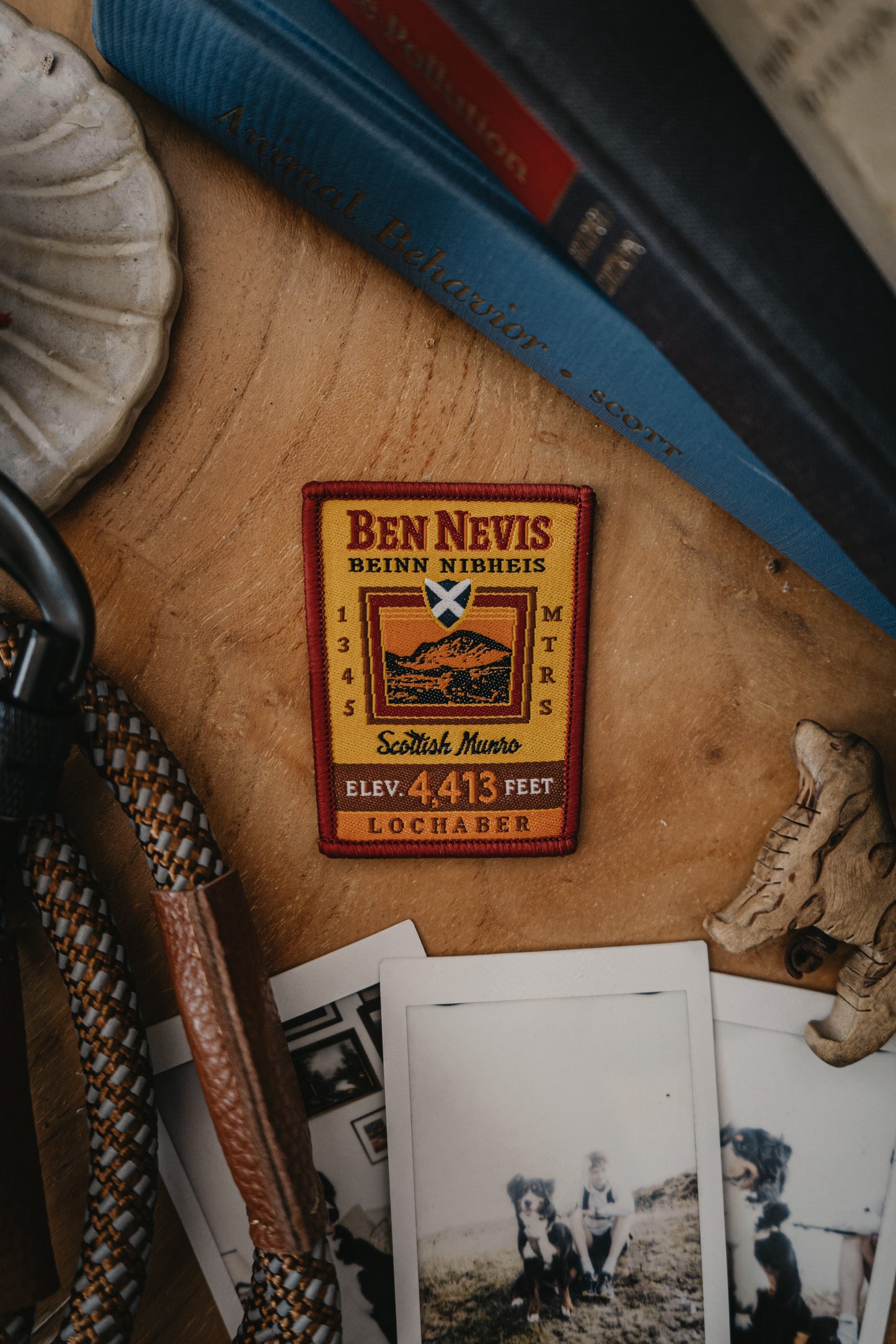 A rectangular embroidered patch reading 'Ben Nevis' with a mountain illustration and elevation 4413 feet, placed on a wooden surface with books, rope, and vintage photos around it.