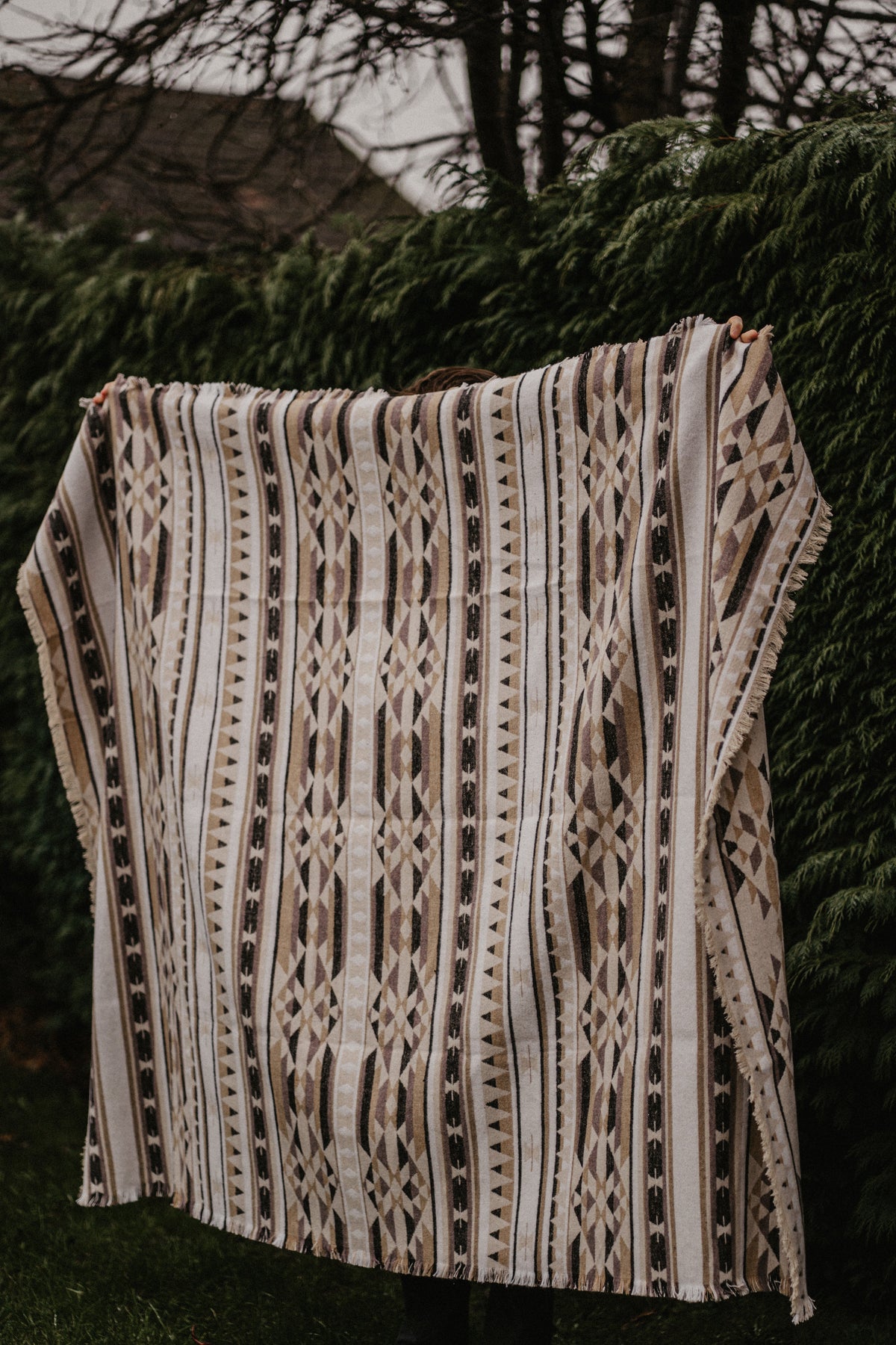 Person holding a patterned woven beige blanket against a hedge
