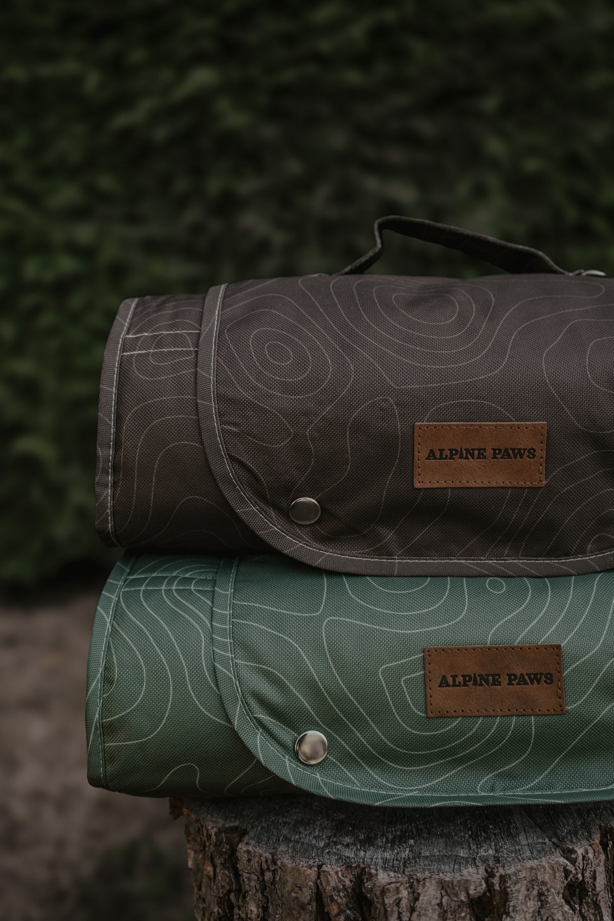 Two rolled travel mats from Alpine Paws stacked on a tree stump, one in dark brown and one in sage green, both with topographic line patterns and brown leather brand patches.