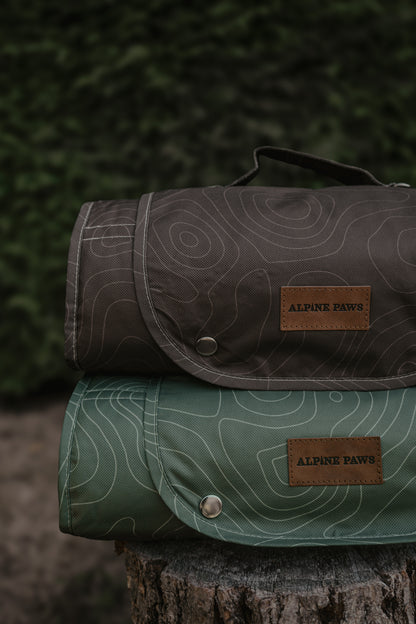Two rolled travel mats from Alpine Paws stacked on a tree stump, one in dark brown and one in sage green, both with topographic line patterns and brown leather brand patches.