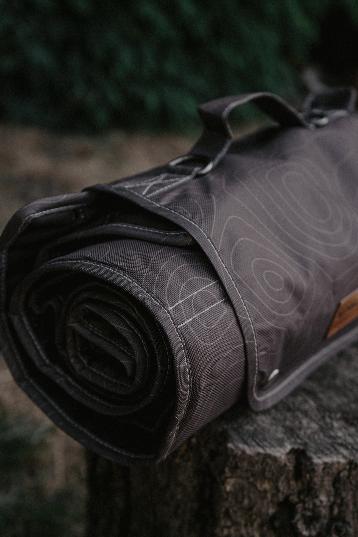 A rolled brown travel mat with a topographic contour line pattern, resting on a rustic tree stump with a blurred forest background.