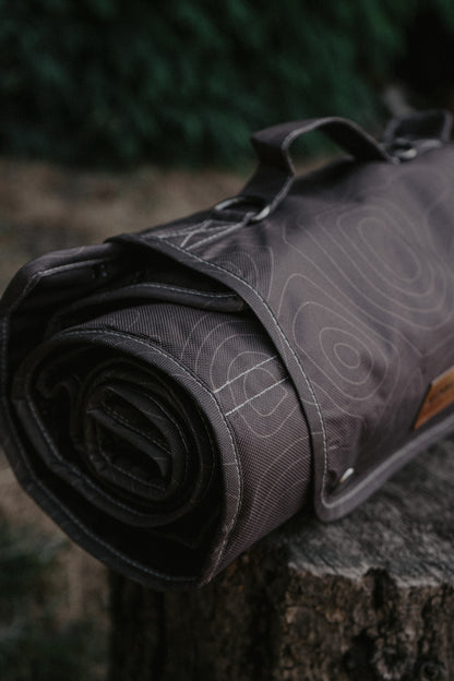 A rolled brown travel mat with a topographic contour line pattern, resting on a rustic tree stump with a blurred forest background.