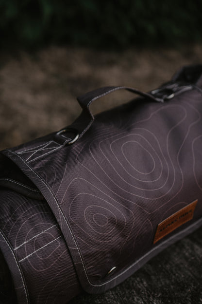 A close-up of rolled travel mat with brown topographic contour line pattern, featuring a brown leather brand patch alpine paws