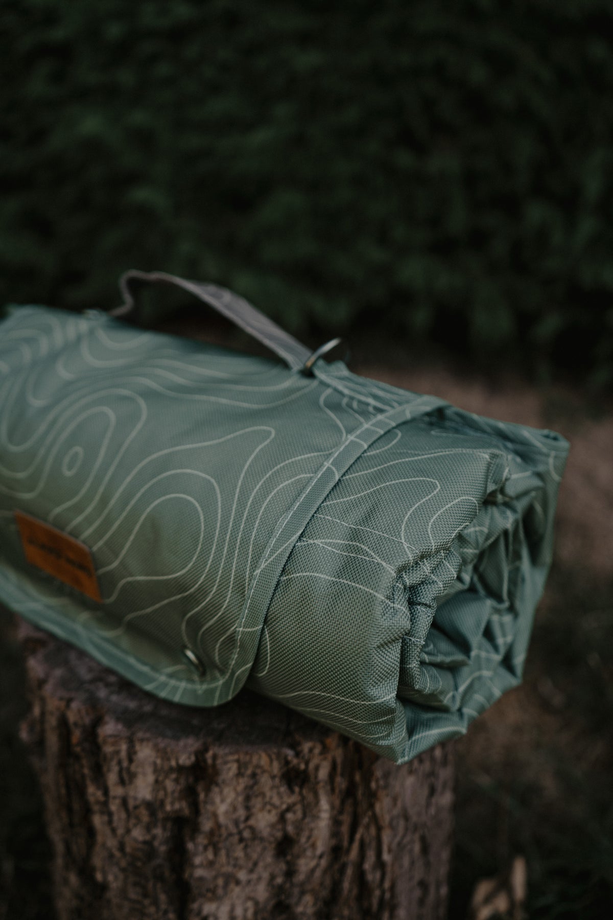 A rolled green settle mat with a white topographic contour line pattern, resting on a rustic tree stump in an outdoor forest setting.