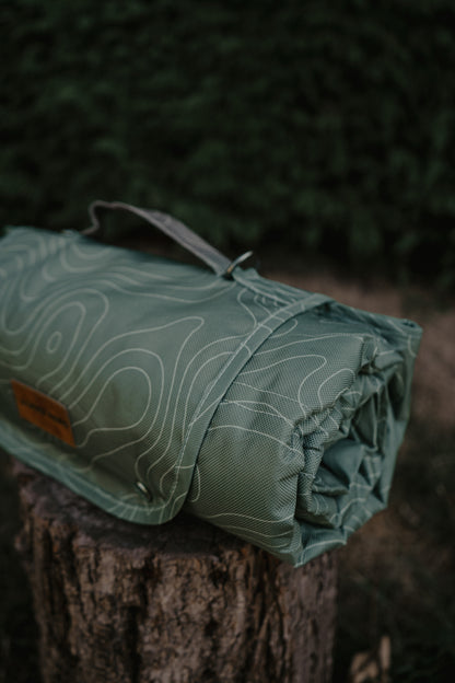 A rolled green settle mat with a white topographic contour line pattern, resting on a rustic tree stump in an outdoor forest setting.