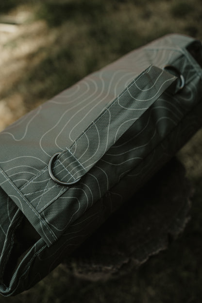 A close-up of a green topographic-patterned dog mat resting on a dark textured surface with a blurred outdoor background.
